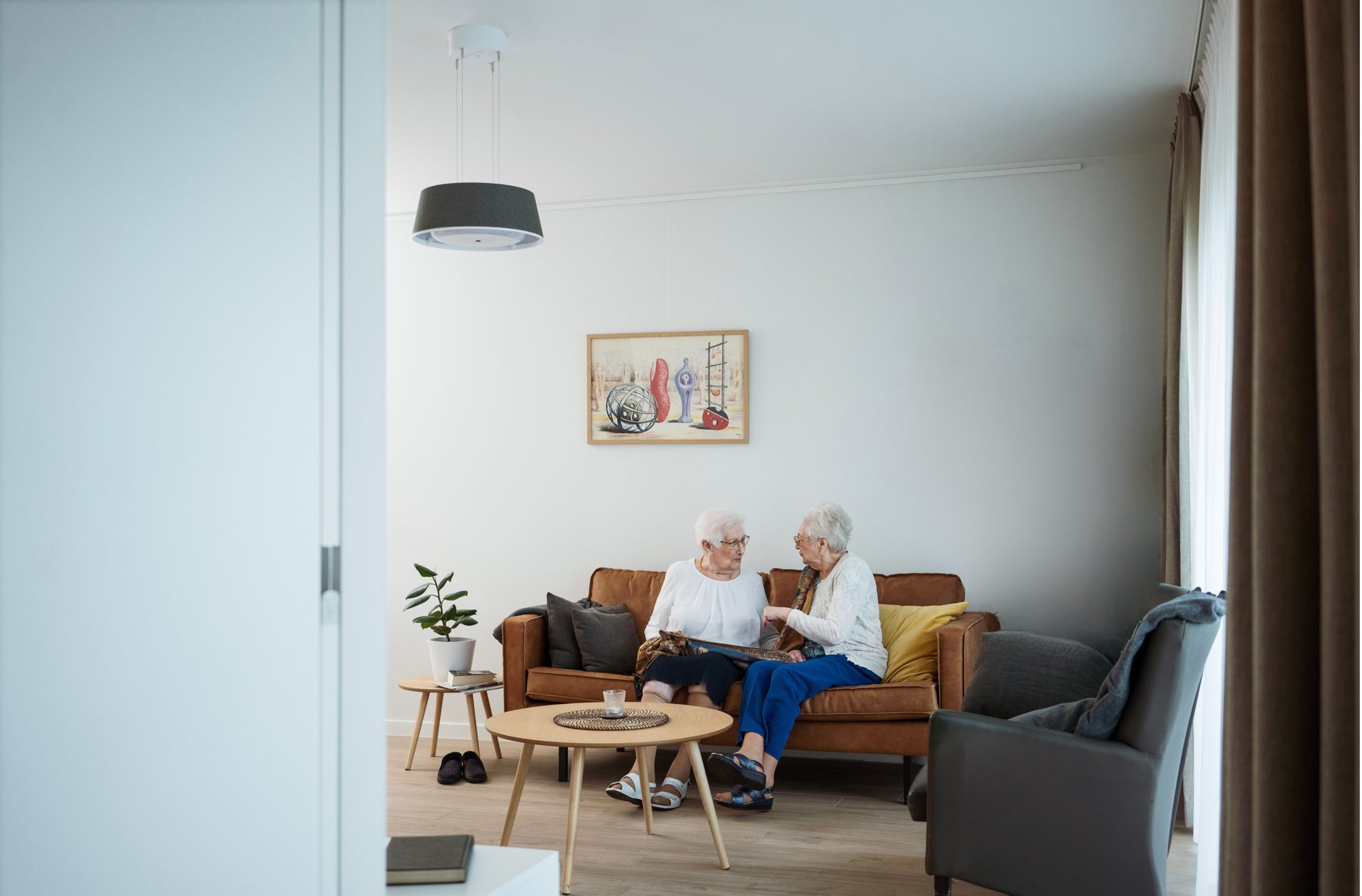 Two residents talking on a sofa in a care home apartment, with a Nobi Pendant light above them.